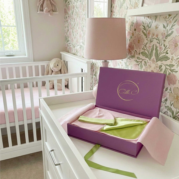 Baby girl nursery, with a purple Alli.C gift box set filled with a pink baby blanket & hat with soft green silk lining sitting on a changing table.